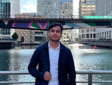 A man wearing a dark jacket over a blue shirt stands on a bridge overlooking a river, with another bridge seen behind him in a city landscape. He is Siddhant Goswami, a data engineer at Yahoo.