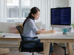 Woman working remotely in a modern office with red brick walls. She is attending a webinar on her computer as there are faces visible on her monitor and she has a notebook and pen on her desk as if she is taking notes.