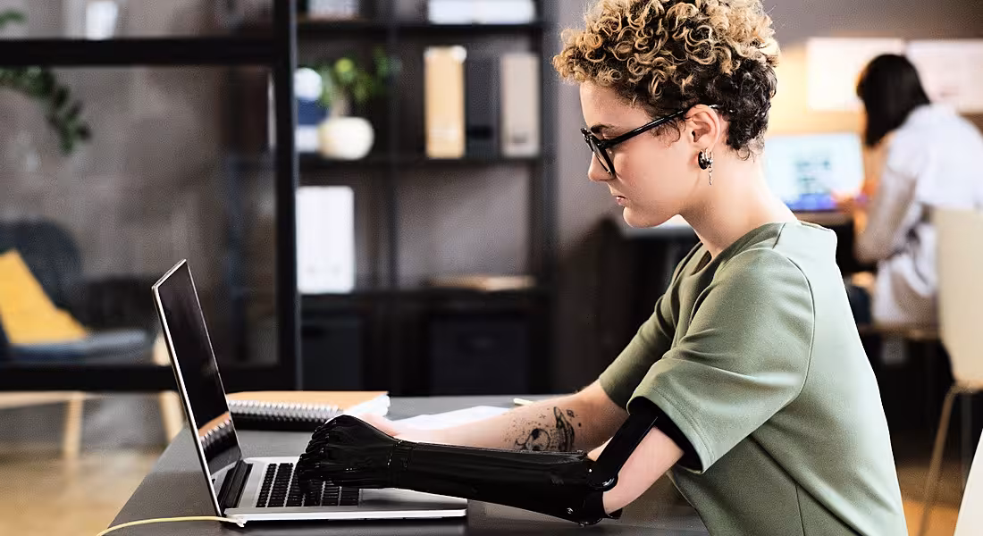 A young professional, with a prosthetic arm, who would benefit from the European Accessibility Act, works at her laptop.