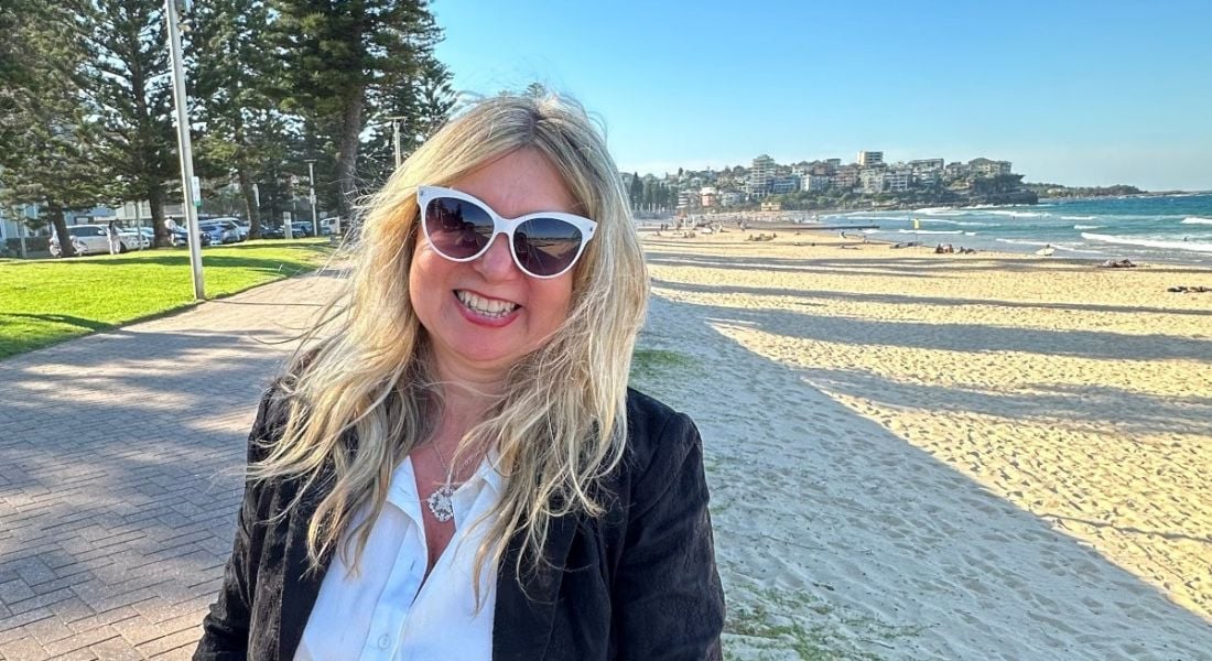 Gamification expert Manolya Kavakli-Thorne. standing outside on a sunny day near a beach smiles at the camera.