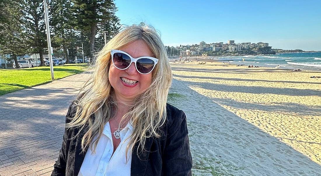 Gamification expert Manolya Kavakli-Thorne. standing outside on a sunny day near a beach smiles at the camera.