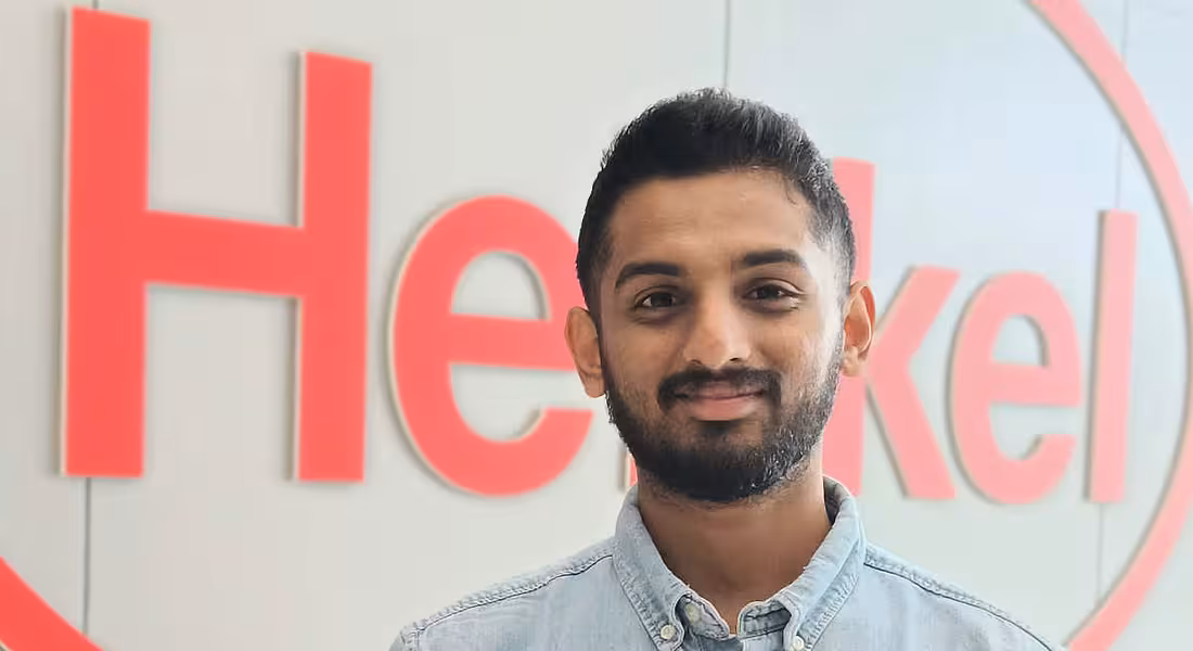 A young man smiles at the camera with the Henkel logo in the background.