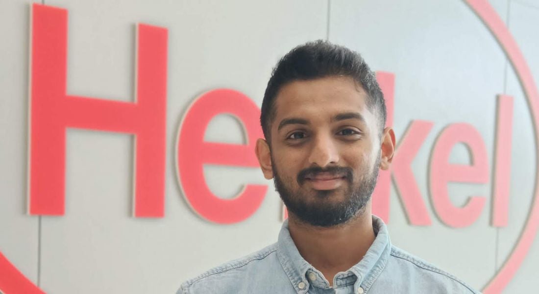 A young man smiles at the camera with the Henkel logo in the background.
