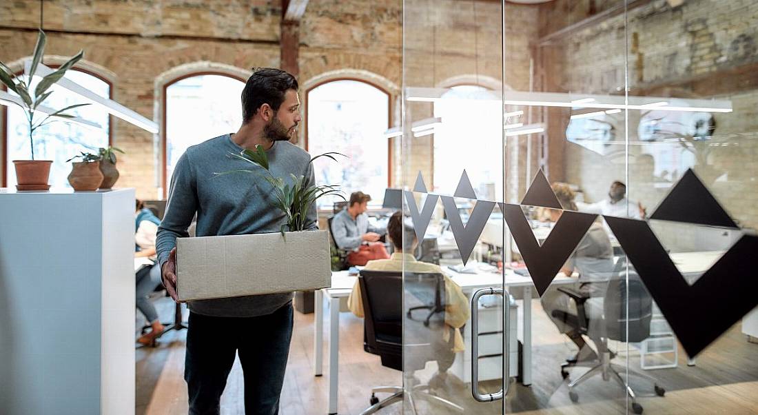 A man carrying a box leaves the office, which has glass walls and people situated throughout, as he has decided to work elsewhere.