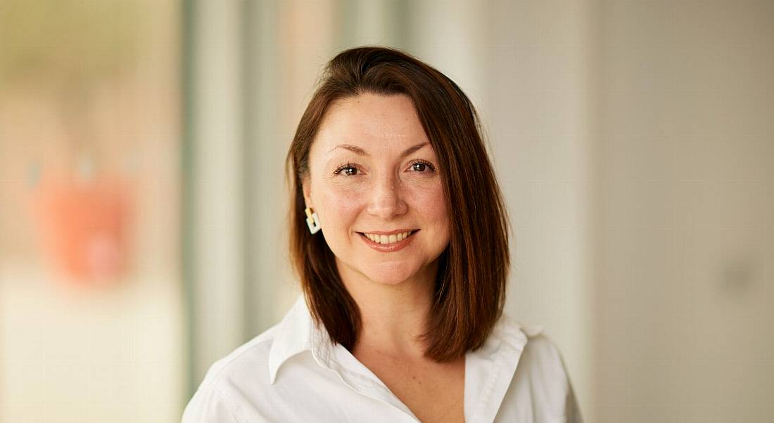 A woman with shoulder-length straight brown hair and a white shirt smiles at the camera in front of a cream-coloured wall.