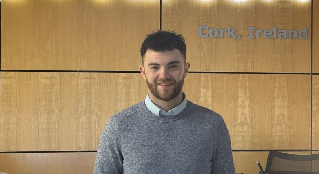 A man with a beard wearing a grey jumper and a white shirt smiles in front of an office wall.