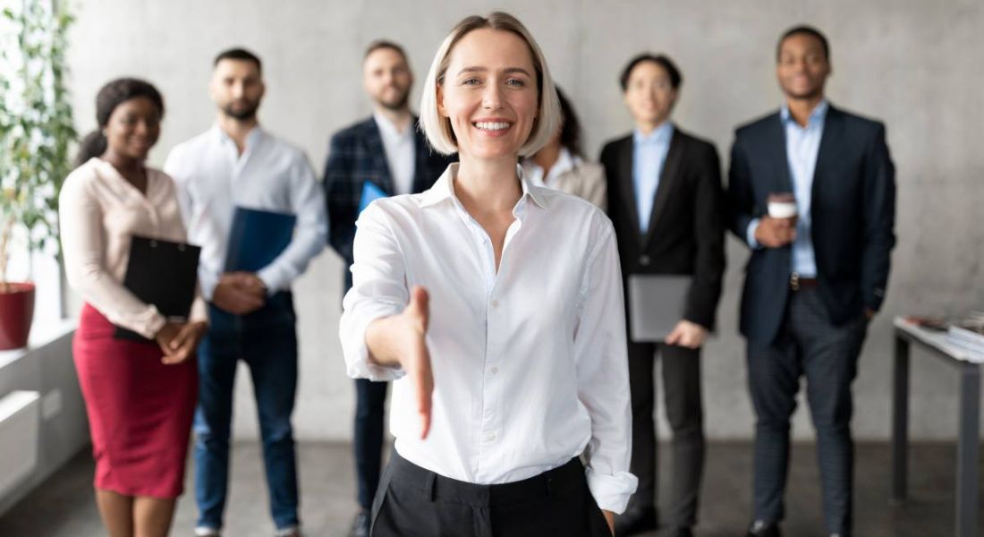 A smiling business woman extends her hand, with her back to her team, indicating a new hire.