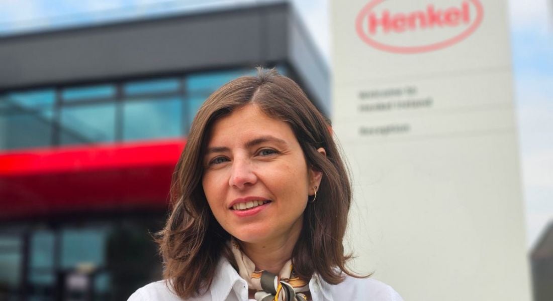 A woman with shoulder-length brown hair smiles in front of an office building and a sign that features Henkel's logo.