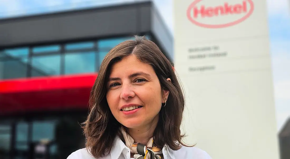 A woman with shoulder-length brown hair smiles in front of an office building and a sign that features Henkel's logo.