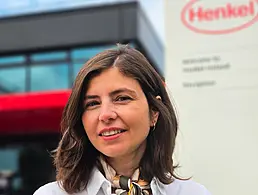 A woman with long, dark hair smiles at the camera in an office. The Henkel logo is on the wall behind her.