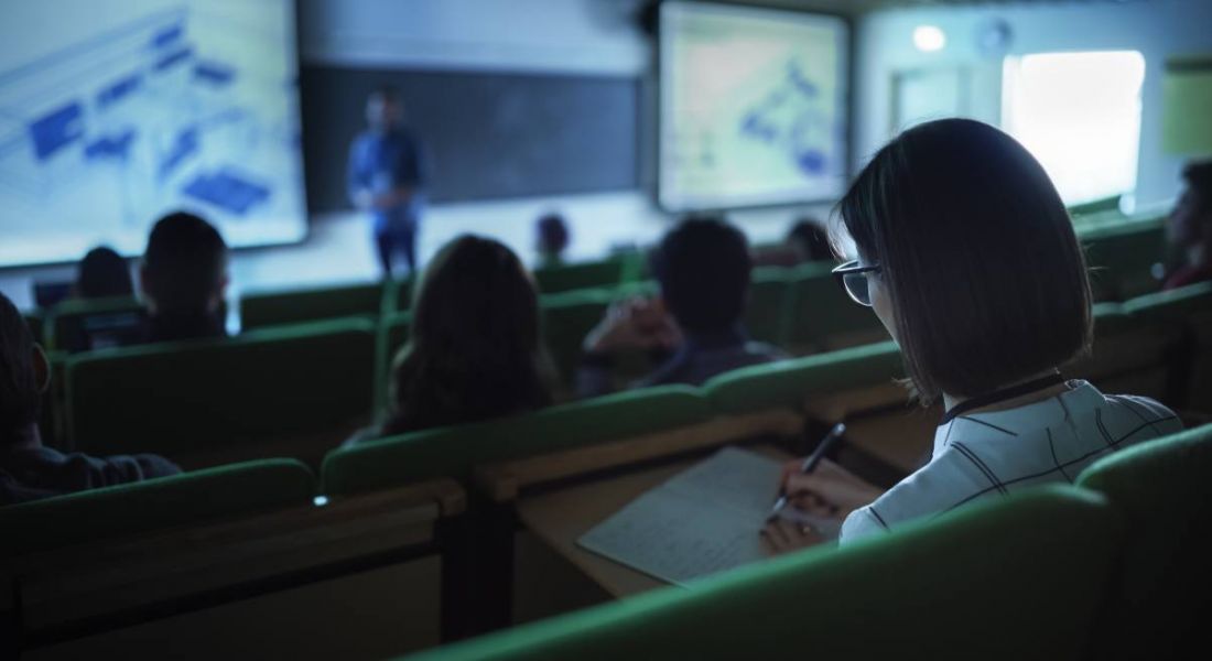 A dark photo of students sitting in a lecture theatre with the lecturer visible at the front of the room below.