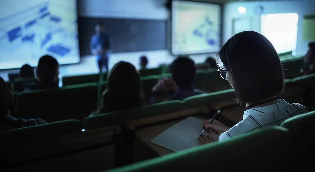 A dark photo of students sitting in a lecture theatre with the lecturer visible at the front of the room below.