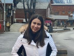 A woman with shoulder-length brown hair smiles in front of an office building and a sign that features Henkel's logo.