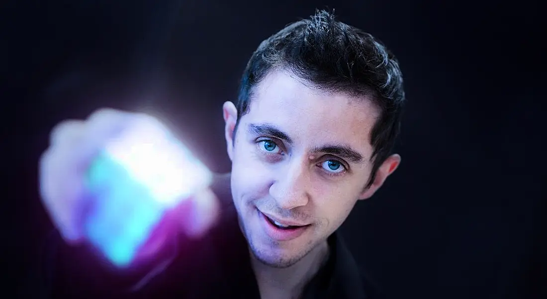 A young man with dark hair looks up at the camera and holds out a completed Rubix cube, which is out of focus.