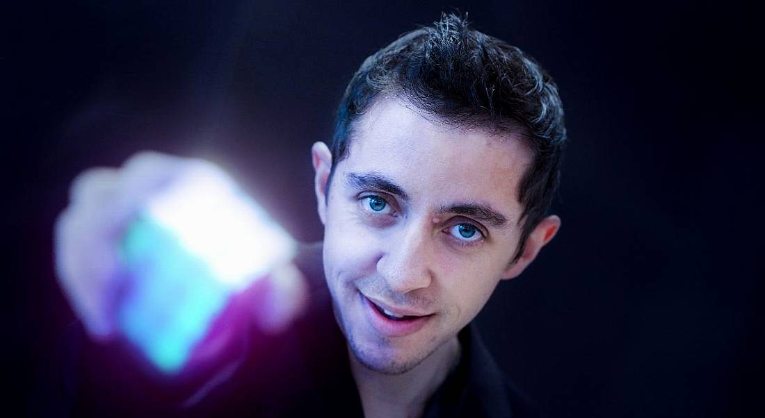 A young man with dark hair looks up at the camera and holds out a completed Rubix cube, which is out of focus.