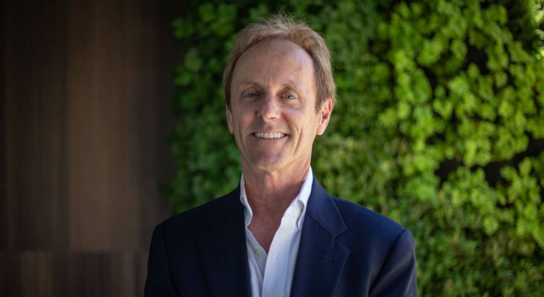 A man wearing a suit jacket and open-neck white shirt smiles at the camera. Behind him is a wall of greenery.