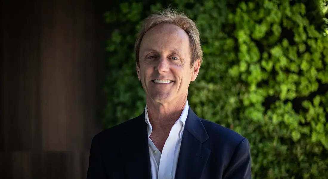 A man wearing a suit jacket and open-neck white shirt smiles at the camera. Behind him is a wall of greenery.