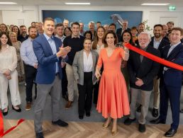 Four people, three men and one woman, at the opening of SaaS company Diligent and its Galway office. They are standing against a wooden panelled wall with balloons in the background.