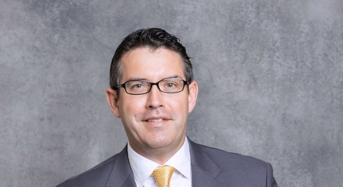 A headshot of a man with dark, short hair and glasses wearing a suit. He is Gary Hanniffy of PwC and he is standing in front of a grey background.