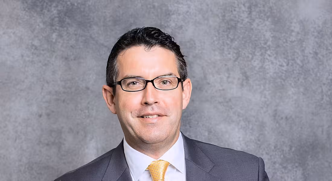 A headshot of a man with dark, short hair and glasses wearing a suit. He is Gary Hanniffy of PwC and he is standing in front of a grey background.