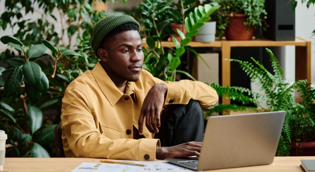 A black man sits at his in-office laptop surrounded by greenery representing being green and sustainable in the workplace.