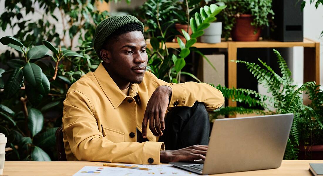 A black man sits at his in-office laptop surrounded by greenery representing being green and sustainable in the workplace.