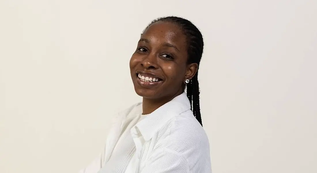 A woman wearing a white shirt smiles at the camera in front of an off-white background.