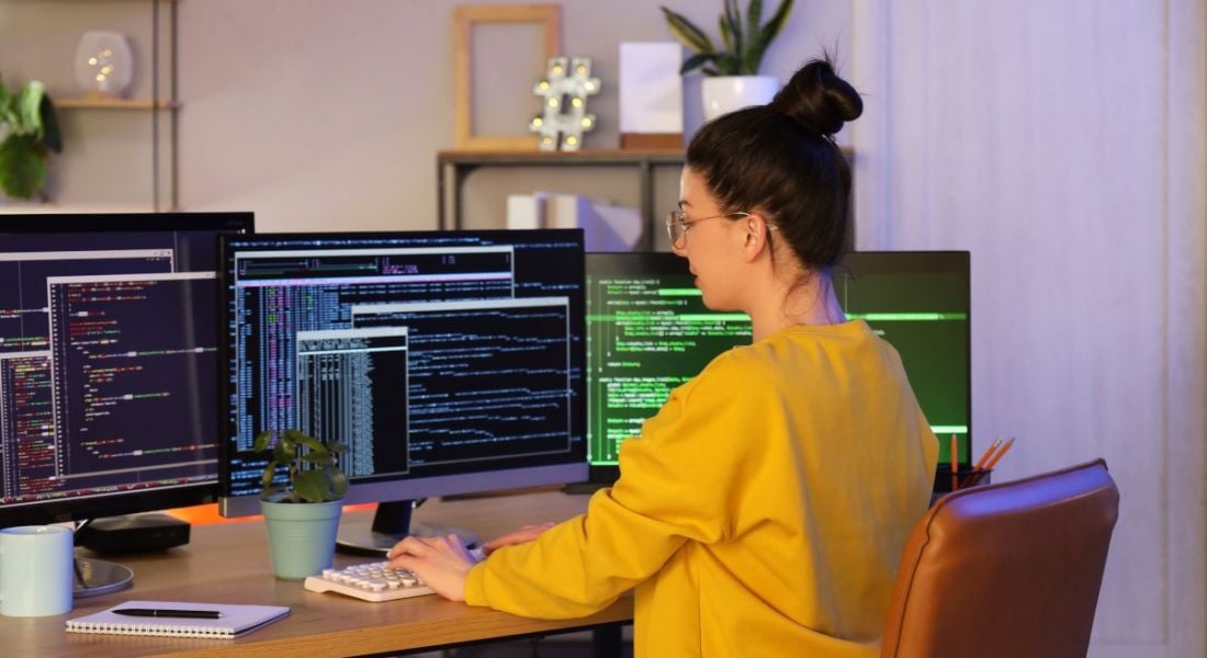 A woman in yellow codes on multiple computer screens in an office setting.