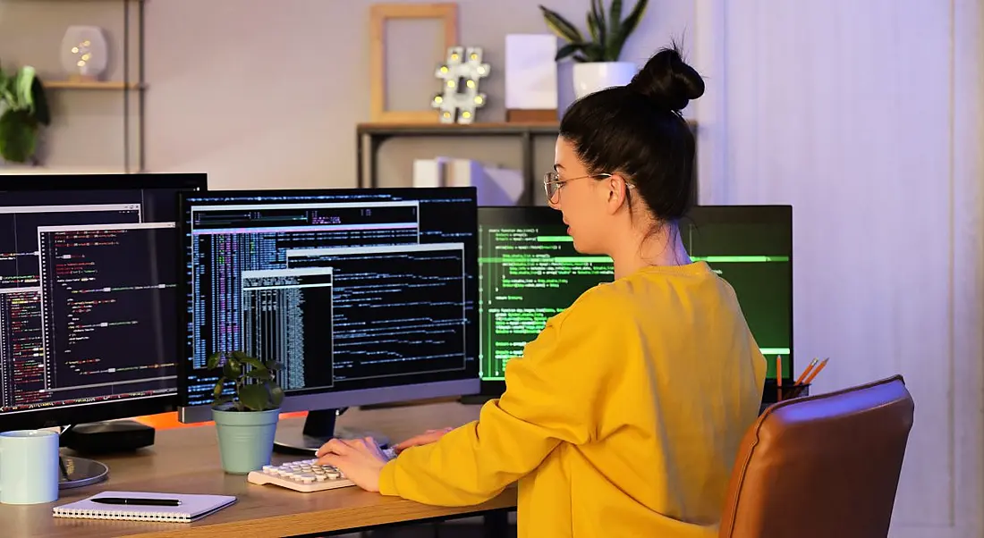 A woman in yellow codes on multiple computer screens in an office setting.