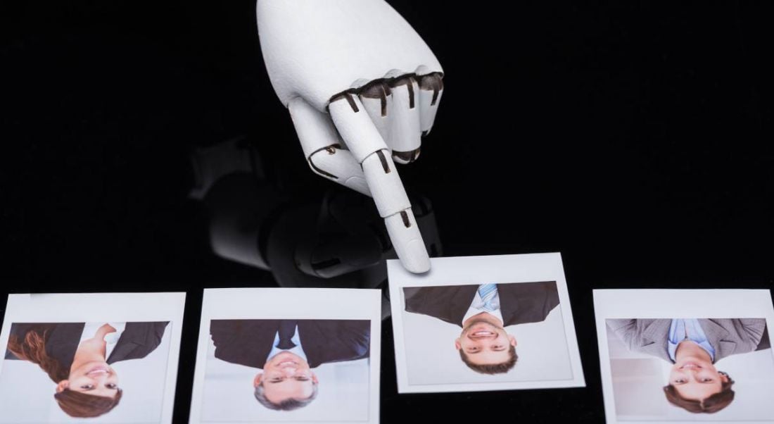 A robotic arm is picking out a male image from a line of job candidates, representing AI in the job application and recruitment process.