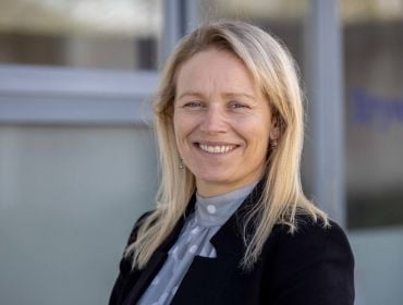 An image of a blonde woman smiling in front of a building. She is Gillian Moody.