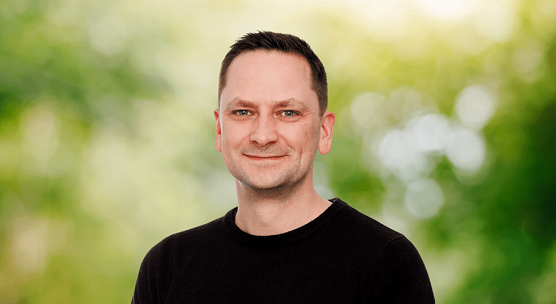 A man wearing a black T-shirt smiles in front of a blurred digital background depicting nature.