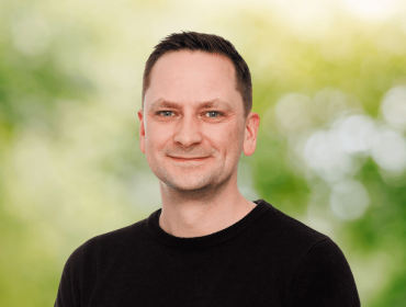 A man wearing a black T-shirt smiles in front of a blurred digital background depicting nature.