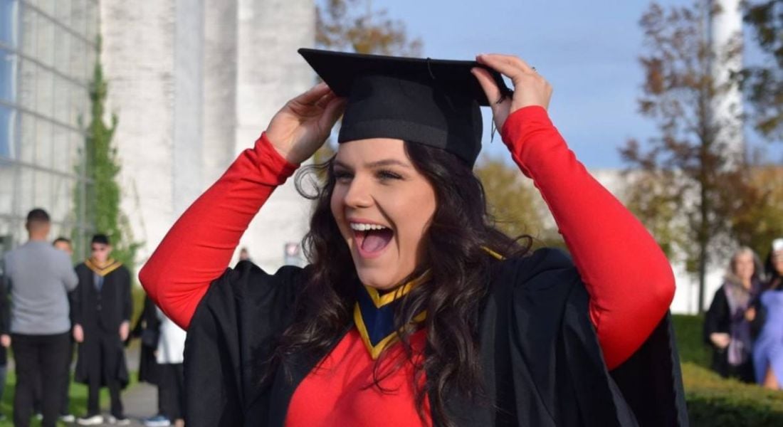 A woman wearing a black graduation gown over a red dress smiles as she she fixes her graduation cap onto her head.