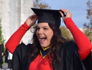 A woman wearing a black graduation gown over a red dress smiles as she she fixes her graduation cap onto her head.