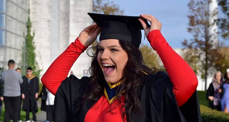 A woman wearing a black graduation gown over a red dress smiles as she she fixes her graduation cap onto her head.