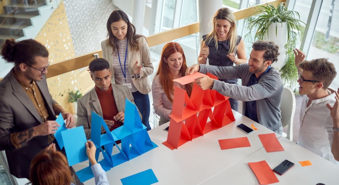 A group of co-workers play a game at work, they are stacking coloured pieces of paper in an activity inspired by workplace gamification.