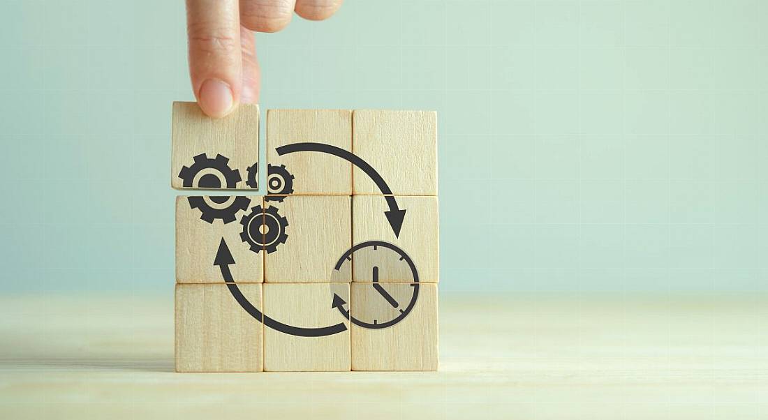 A hand fixes a final wooden block to reveal a number of cogs working towards the better optimisation of time.