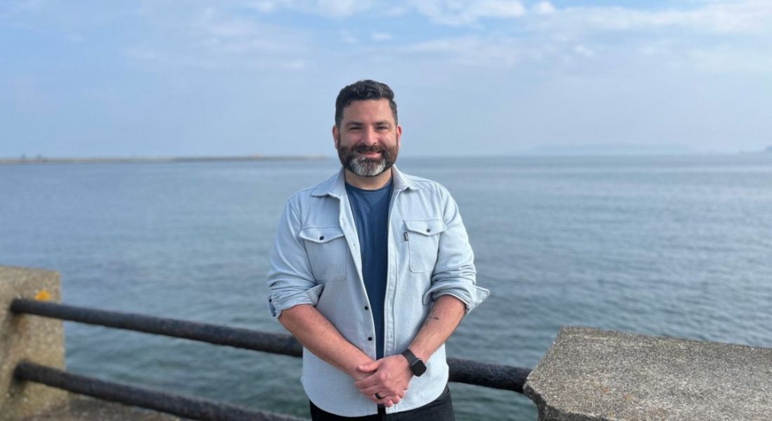 A man wearing a light blue shirt with rolled up sleeves smiles while standing in front of the sea.