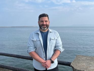 A man wearing a light blue shirt with rolled up sleeves smiles while standing in front of the sea.