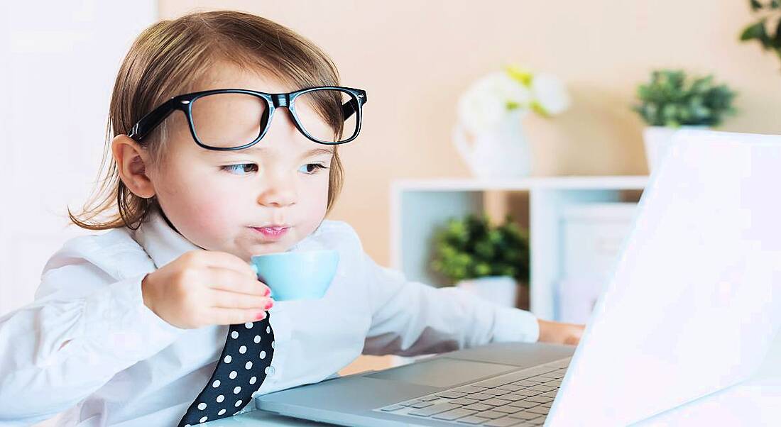 A toddler with glasses, a tie and a cup of coffee sits at a laptop doing a day's work.