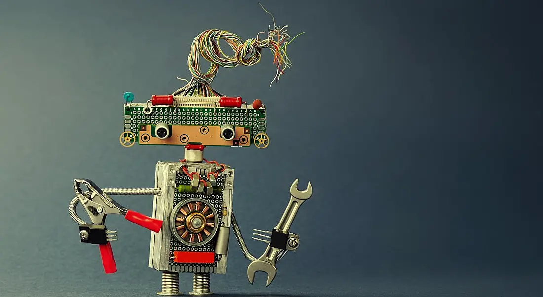 A metal toy robot made up of various electrical components. It is holding a wrench and pliers.