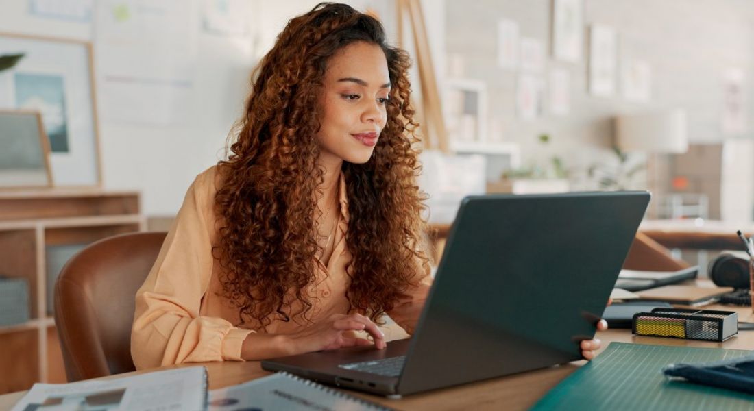 A young woman with long brown hair working on a laptop with a scattering of stationery around her. She symbolises a tech contractor.