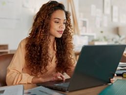 A woman is working at a laptop in a bright office space and smiling.