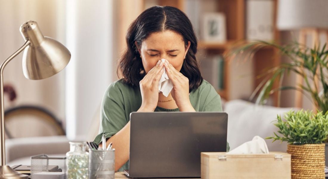 A sick woman sits at her laptop, blowing her nose and trying to work.