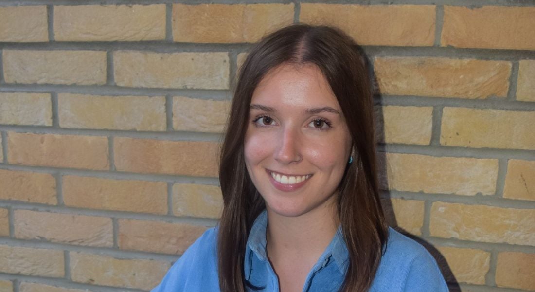 A woman with long brown hair wearing a blue shirt smiles while sitting in front of a brick wall.