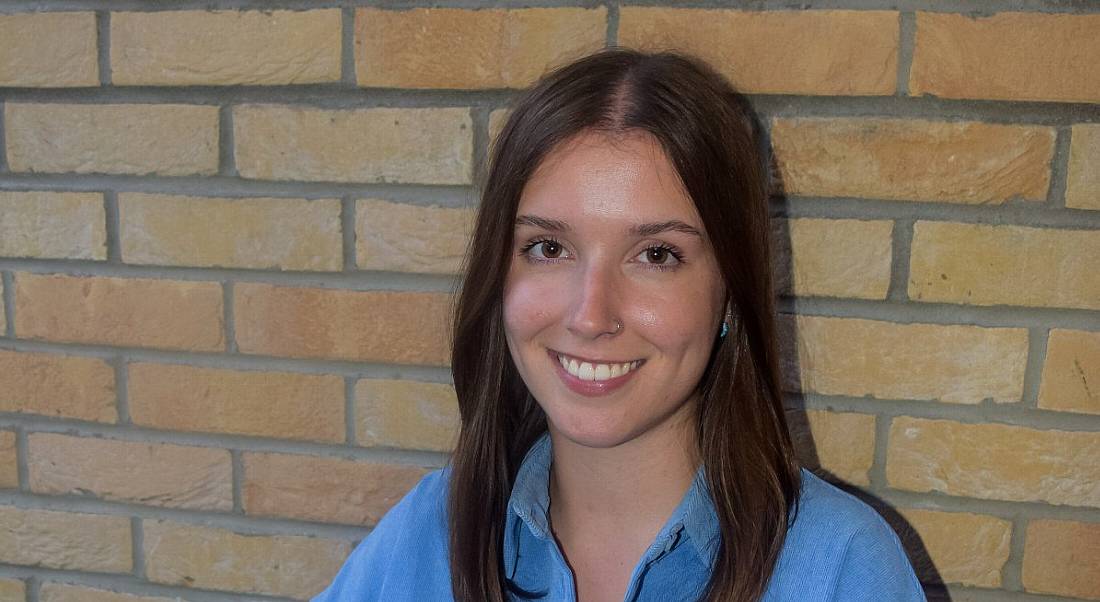 A woman with long brown hair wearing a blue shirt smiles while sitting in front of a brick wall.