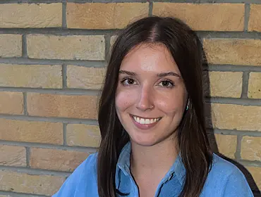 A woman with long brown hair wearing a blue shirt smiles while sitting in front of a brick wall.