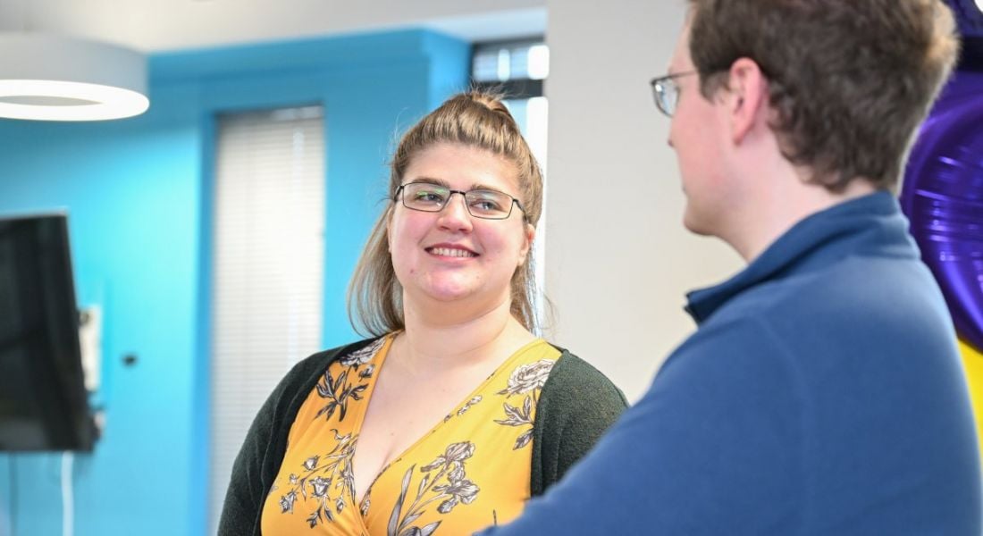 A woman wearing a yellow patterned blouse and glasses smiles while talking to a colleague to the right of the image.