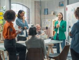 Woman in glasses, with a top bun, in yellow, is shushing, indicating what not to say.